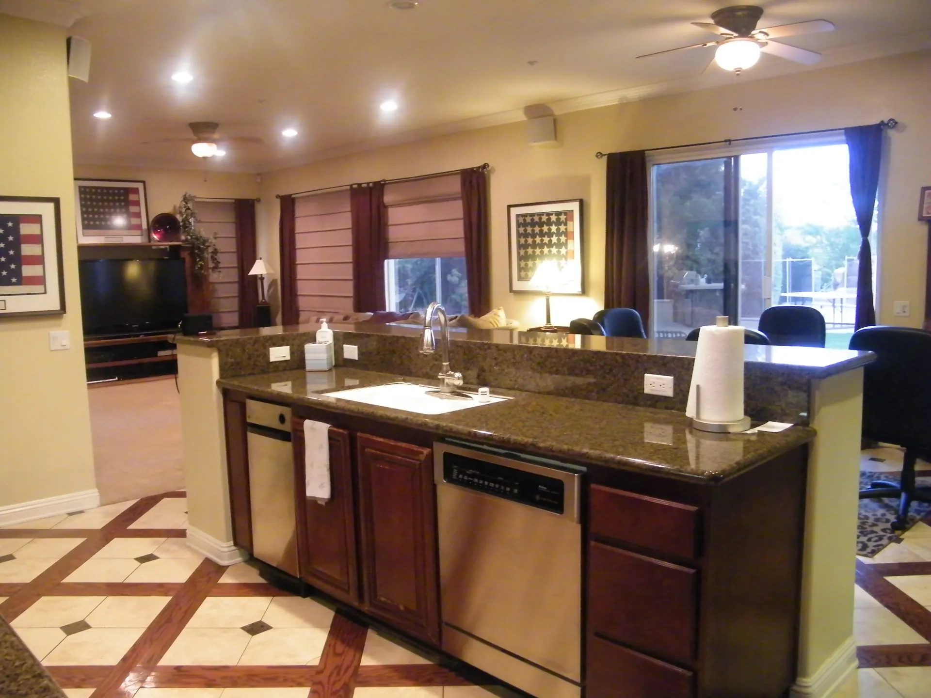 An open kitchen in a single-family home, with a granite-topped island holding the sink, a stainless steel dishwasher and dark wood cabinets, looking toward the family room and a sliding glass door to the backyard.