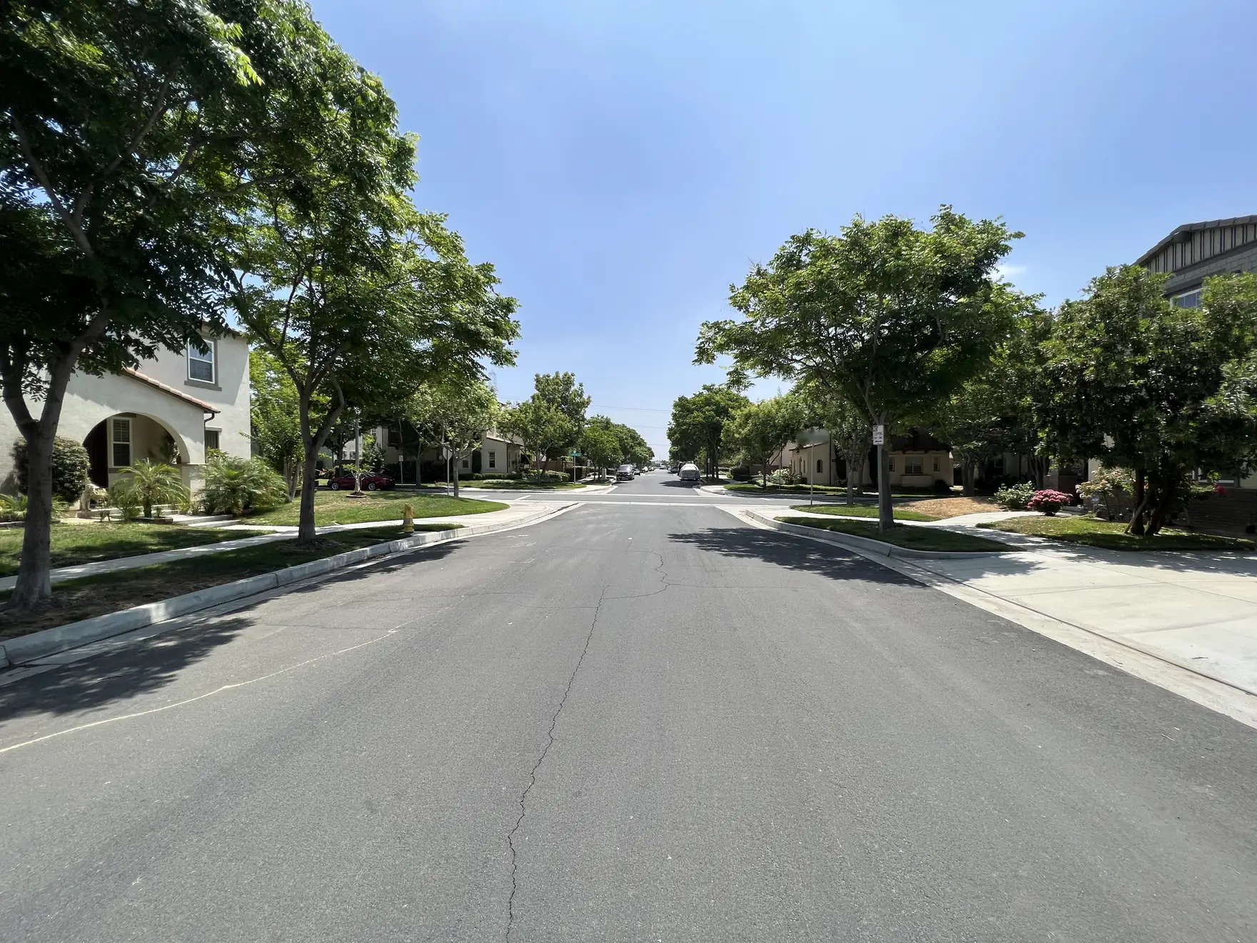 A quiet, tree-lined residential street with two-story stucco homes and green front lawns on a sunny day.