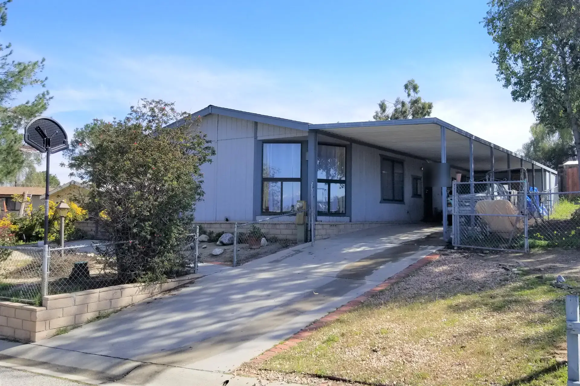 A single-story blue-gray manufactured home with a long metal carport, chain-link fencing, a low block wall and a basketball hoop by the driveway, under a clear blue sky.