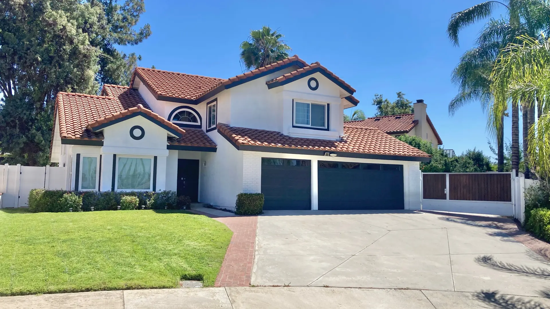 Two-story stucco house with a red tile roof, a three-car garage and an empty driveway, palm trees along the street.