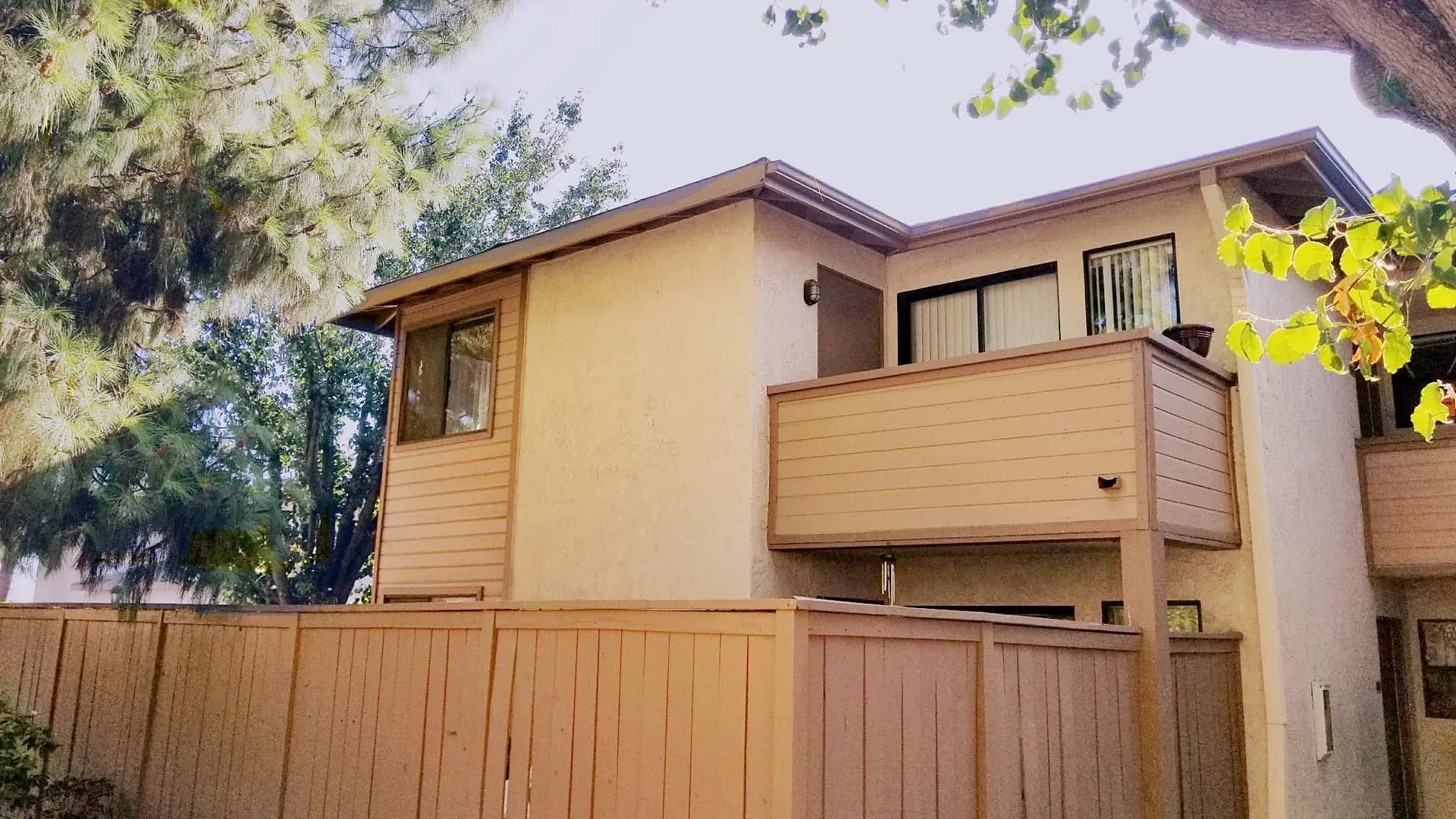 Two-story attached townhome with a second-floor balcony and vertical blinds in the windows, behind a wood privacy fence, with pine and sycamore branches overhead.