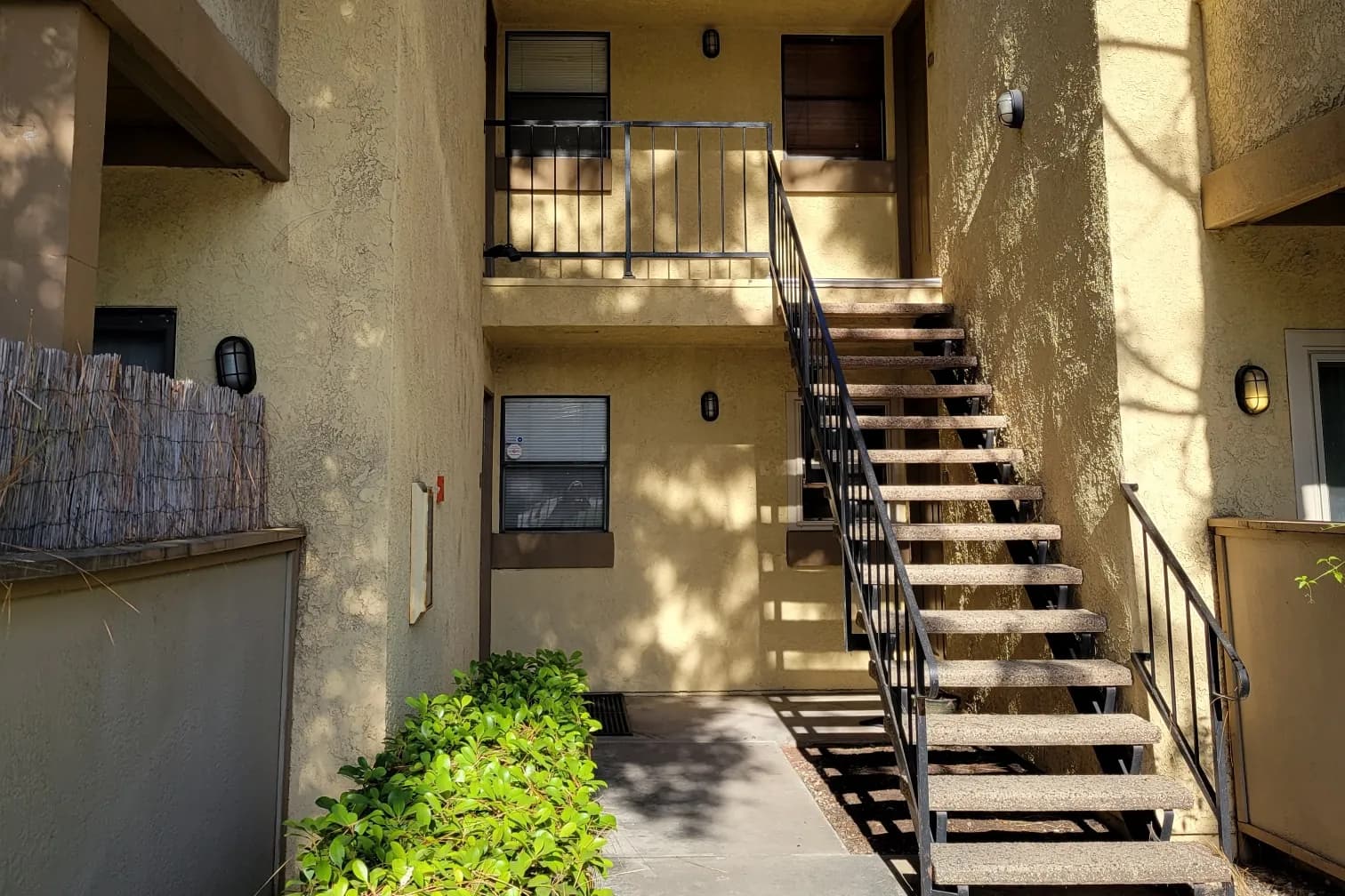 An exterior metal staircase leading to a second-floor unit in a two-story stucco apartment building with tile roof trim, wall lanterns and a hedge along the walkway.