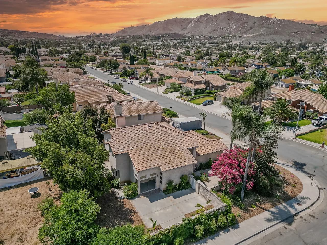 Aerial view of a neighborhood of tile-roofed single-family homes along curving residential streets, with hills under an orange sunset sky.
